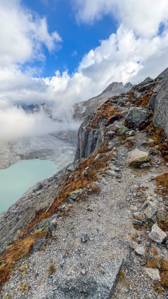 Rocky trail with autumn grass above the Rhône Glacier lake, fog in the valley