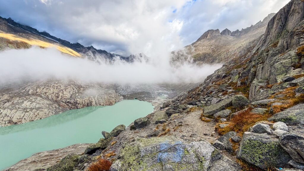 Rocky trail with a blue paint mark above the Rhône Glacier lake, fog drifting over the glacier